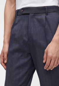 Gros plan d'une personne portant un pantalon plissé gris foncé et une chemise blanche, montrant les mains reposant sur les côtés.