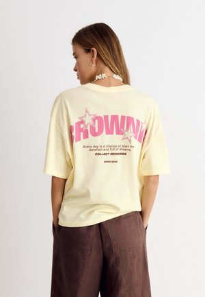 Femme aux longs cheveux porte un T-shirt oversize jaune clair avec un texte rose et des étoiles dans le dos, ainsi qu'un pantalon marron, debout les mains dans les poches.