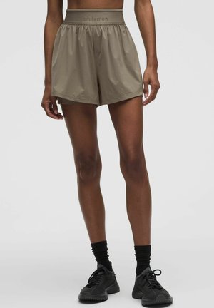 Persona con pantalones cortos Lululemon de color marrón claro, calcetines negros y zapatos deportivos negros sobre un fondo gris liso.