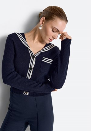 Žena v tmavomodrom kardigane s bielymi pruhmi a perlovými náušnicami, pózujúca so sklonenou hlavou a rukou pri čele na pozadí bielej steny.