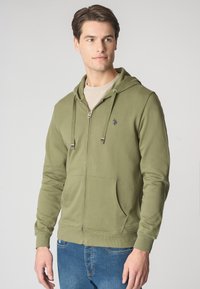 Felpa verde con zip, con tasca frontale, lacci e logo. Tessuto morbido con polsini e orlo a costine. Indossata sopra una t-shirt beige e jeans blu.