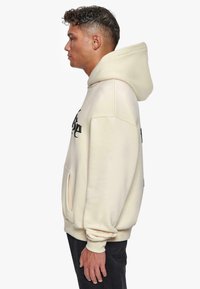 Cremefarbener Hoodie mit einer geräumigen Kapuze, Fronttasche und schwarzem Textdesign. Weicher, strukturiertes Material, enge Bündchen und lässige Silhouette.