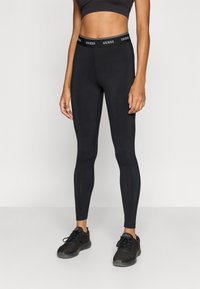 Svarte leggings laget av elastisk materiale, med tettsittende design og en elastisk midjebånd med "GUESS"-logo trykt langs toppen.