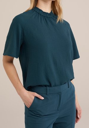 Türkisfarbenes Kurzarmtop mit gerafftem Ausschnitt, strukturierter Stoff, lockere Passform, kombiniert mit passenden, maßgeschneiderten Hosen, Hand in der Tasche.