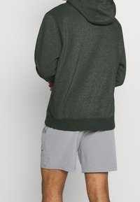 Tmavě zelená mikina s kapucí vyrobená z texturované látky s tkaničkou. Nosená přes světle šedé šortky se bočními kapsami a malým logem.