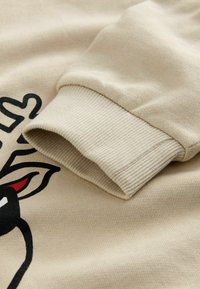 Manica di felpa beige con polsino a costine, che mostra parte di una grafica di volto di animale nero e rosso sul tessuto.