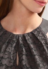 Robe en dentelle noire avec des motifs floraux complexes, design plissé au niveau du col, et accents en satin sur les épaules. Détail en forme de clé à l'avant.