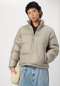 Beige puffer jacket with a high collar, zip closure, and stitched panels. Worn over a dark shirt, held with one hand, paired with light denim.