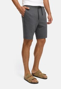Hombre vestido con pantalones cortos grises oscuros con cordón y sandalias beige, de pie con una mano en el bolsillo sobre un fondo blanco.