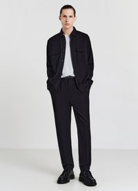 Camicia nera con bottoni e due tasche sul petto sopra una maglietta grigia chiara, abbinata a pantaloni neri e scarpe nere. Tessuti dal aspetto liscio.