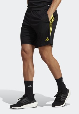 Mann trägt schwarze Sportshorts mit gelben Streifen, schwarze Adidas-Socken und schwarze Laufschuhe, steht mit einer Hand in der Tasche.
