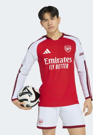 Junger männlicher Fußballspieler hält einen schwarz-weißen Ball, trägt rote und weiße Arsenal-Trikot mit dem Logo "Emirates Fly Better".