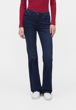 Person trägt dunkelblaue Straight-Leg-Jeans, weiße Sneaker und ein rotes Langarmshirt und steht vor einem einfarbigen Hintergrund.