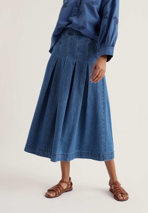 Vrouw in een geplooide denim midi-rok, blauwe blouse met lange mouwen en borduursel, en bruine lederen platte sandalen, staand tegen een effen achtergrond.