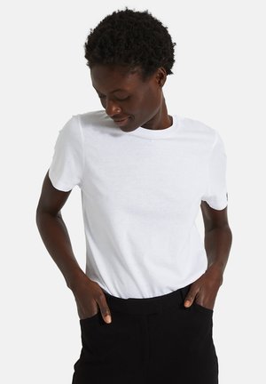 T-shirt blanc en coton à manches courtes avec un col rond. Porté avec les mains dans les poches, associé à un bas noir, mettant en avant un design simple.