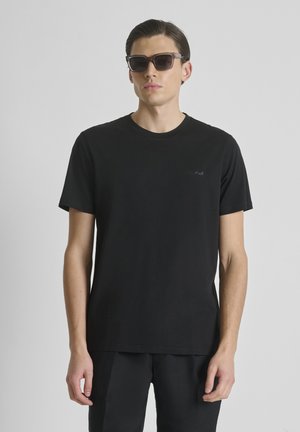 Uomo con t-shirt nera a maniche corte e pantaloni neri, con occhiali da sole scuri, in piedi contro uno sfondo chiaro e uniforme.