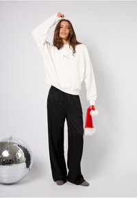 Felpa bianca oversize con ricami rosa, abbinata a pantaloni neri a gamba larga con stampa leopardata. Il modello tiene in mano un cappello da Babbo Natale rosso con rifiniture bianche.