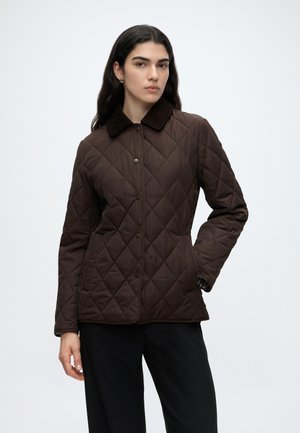 Femme aux longs cheveux foncés portant une veste matelassée marron avec un col foncé et un pantalon noir, debout avec une main dans la poche.