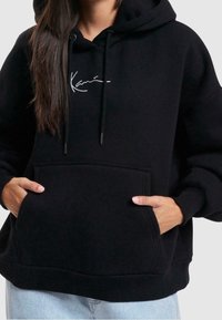 Felpa nera con cappuccio realizzata in tessuto morbido, dotata di una tasca a marsupio frontale, cordini e un piccolo logo bianco sul petto. Vestibilità rilassata.