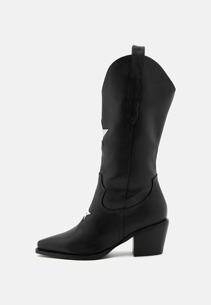 Musta nahkasaapas cowboy-tyylillä, jossa on terävä kärki, keskipolven korkeus, hopeiset tähtiakoristeet ja paksu korko. Saumojen yksityiskohdat ovat näkyvissä.