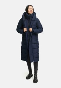 Palton lung de tip puffer de culoare bleumarin, cu guler înalt, design matlasat, închidere cu fermoar în față și buzunare laterale. Asortat cu pantaloni negri și cizme.