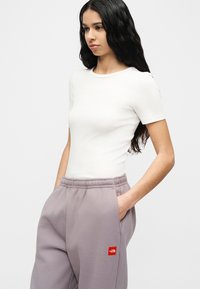 Γυναίκα με μακριά σκούρα μαλλιά που φοράει εφαρμοστή λευκή μπλούζα με κοντά μανίκια και ανοιχτόμωβ φόρμα με κόκκινο λογ ότυπο.