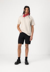 Beige t-skjorte, svarte shorts, svarte klumpete sko. Rød bandana rundt halsen. Modell som står med den ene hånden i lommen, nøytral bakgrunn.