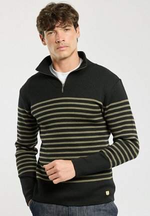 Mand med kort brunt hår iført sort kvart lynlås sweater med olivengrønne vandrette striber og mørke jeans mod en ensfarvet baggrund.
