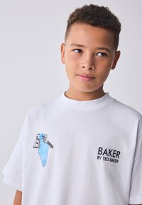 Berniukas su trumpais garbanotais plaukais, vilkintis baltą marškinėlį su mėlynu raktų pakabuku ir užrašu „BAKER BY TED BAKER“ ant vienspalvio fono.