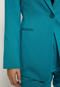 Tīkamais blazera dizains ir ar vienu pogu, sānu kabatām un strukturētu piegriezumu. Izgatavots no gludas auduma ar viegli pamanāmu tekstūru.