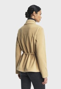 Veste en polaire beige à taille cintrée, avec un col à revers et des manches longues, sur un fond blanc uni.