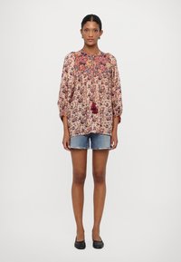 Langärmlige Bluse mit Blumenmuster in Hellrosa, mit filigraner Stickerei und Quastendetails, kombiniert mit Jeansshorts und schwarzen Ballerinas.