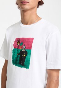 Hvid bomuld t-shirt med et farverigt grafisk design af to figurer, den ene med en spids hat og den anden i en sort frakke, på en tekstureret baggrund.
