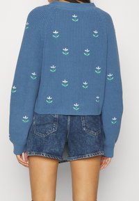 Pull en tricot bleu avec broderie florale en blanc et vert. Design court avec des manches longues et texture côtelée, associé à un short en denim.