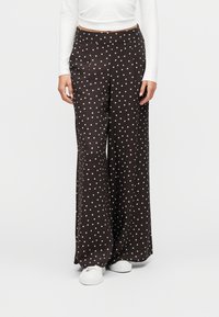 Γυναίκα φοράει μαύρο παντελόνι φαρδιά γραμμή με λευκές πουά, λευκό μακρυμάνικο μπλουζάκι και λευκά αθλητικά, στέκεται μπροστά σε ένα απλό φόντο.