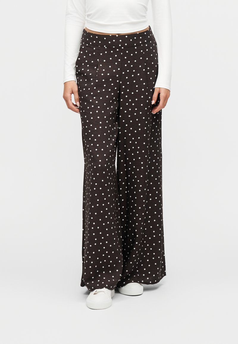 Γυναίκα φοράει μαύρο παντελόνι φαρδιά γραμμή με λευκές πουά, λευκό μακρυμάνικο μπλουζάκι και λευκά αθλητικά, στέκεται μπροστά σε ένα απλό φόντο.