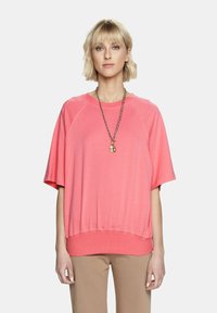 Korallowa różowa bluzka z krótkim rękawem o luźnym kroju i prążkowanym wykończeniu. W komplecie z beżowymi spodniami i długim naszyjnikiem z zawieszką.