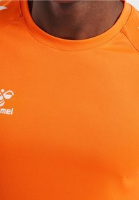 Camiseta deportiva de color naranja brillante, hecha de una tela suave y que absorbe la humedad. Presenta un escote redondo y un logo en el lado izquierdo del pecho.