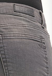 Jeans de denim gris con una etiqueta en la cinturilla. Las características incluyen bolsillos traseros, costuras finas y una textura suave. La tela parece ligeramente desgastada.