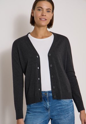 Femme portant un cardigan gris foncé boutonné par-dessus une chemise blanche et un jean bleu, debout devant un fond clair uni.