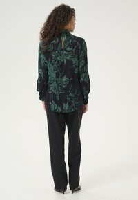 Vista posteriore di una blusa floreale con motivi verdi su sfondo nero, caratterizzata da un alto collo e dettaglio a chiave, abbinata a pantaloni neri.