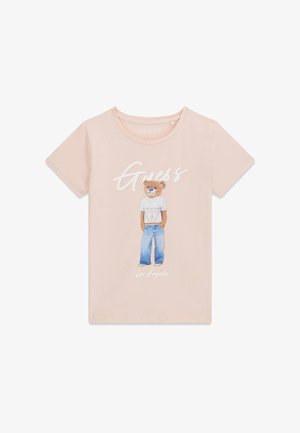 Hellrosa T-Shirt mit einem Grafikdruck eines Bären, der ein weißes Guess-T-Shirt und blaue Jeans trägt, mit dem Schriftzug „Guess“ darüber.