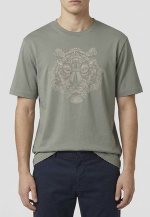 Hombre con camiseta verde oliva con un tigre bordado detalladamente en el centro del pecho, combinado con pantalones azul marino oscuro.
