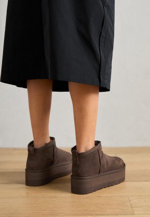 Personne portant une jupe foncée au genou et des bottines à plateforme en daim marron, debout sur un sol en bois.