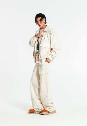 Joven con rastas cortas que lleva una chaqueta y pantalones crema extragrandes con puntos reflectantes, camiseta gráfica y botas de trabajo marrones, posando de lado.