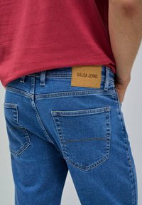 Jeans in denim blu con una toppa in pelle beige su cui è scritto "SALSA JEANS", due tasche posteriori con dettagli di cucitura e una vestibilità a vita media.