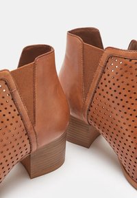 Brązowe skórzane botki za kostkę z wycięciami perforacji, elastycznymi panelami bocznymi oraz niskim, ułożonym drewnianym obcasem. Gładka tekstura w całości.