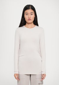 Jeune femme aux longs cheveux noirs portant un haut ajusté à manches longues beige clair et un pantalon assorti, se tenant devant un fond blanc uni.