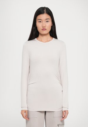 Ung kvinne med langt, svart hår iført lysebeige langermet tettsittende topp og matchende bukser, står mot en ensfarget hvit bakgrunn.