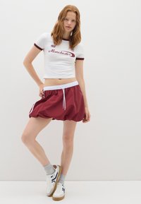 Joven mujer con cabello rojo, vistiendo una camiseta corta blanca con el texto "Manchester" y pantalones cortos deportivos color burdeos, de pie frente a un fondo blanco liso.